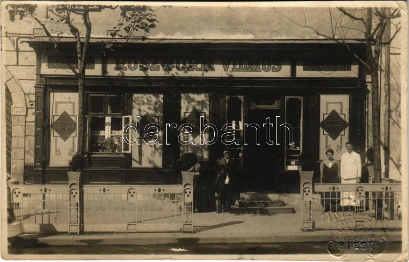 1924 Budapest I. Ruszwurm Vilmos cukrászdája. Szentháromság utca 7. Diskay photo (fa)  A budai várban, a Szentháromság utca 7. számú házban 1827-ben kezdte meg működését a Ruszwurm cukrászda elődje Schwabl Ferenc cukrász vezetésével. 1884-ben az addig segédként dolgozó, s a családba beházasodó Ruszwurm Vilmos vette át a cukrászda vezetését, amelynek egészen 1922-ig állt az élén.  A Ruszwurm Cukrászda belső tere klasszikus, 19. századi polgári hangulatot áraszt, sötét fabútorokkal és patinás vitrinek­kel. Az időtlen berendezés és a meghitt, nosztalgikus atmoszféra hűen tükrözi Budapest egyik legrégebbi cukrászdájának történelmi múltját.  A képeslapon a cukrászda 1924-es formája látható: az ablakot és a bejáratot a maitól eltérően egy előtérré, kirakattá alakították, üvegszekrényében cukorkakínálók és asztaldíszek gyűjteménye kapott helyett. A képeslapra írt üzenet angol nyelvű szövegében a következő olvasható: "Ez az a picike Viktória előtti időből származó hely, ahol kávét ittunk."  A bejárat előtt maga Ruszwurm Vilmos balról a második alak, sapkában, jellegzetes módon, hátratett kézzel áll. A fotólap 1924-ben készült, tehát az idős Ruszwurm ekkor már két éve hivatalosan visszavonult, a vezetést társára Tóth Ferencre bízva, aki a patinás hely iránti tiszteletből a nevet változatlanul hagyta.  A cukrászdát 2025-ben határozatlan időre bezárták.  //  In the Buda Castle, at Szentháromság Street 7, the Ruszwurm Confectionery began operating in 1827 under the leadership of its predecessor, confectioner Ferenc Schwabl. In 1884, Vilmos Ruszwurm, who had previously worked as an assistant and married into the family, took over the management of the confectionery, which he headed until 1922.  The interior of the Ruszwurm Confectionery exudes a classic, 19th-century bourgeois atmosphere, with dark wooden furniture and patinated display cases. The timeless furnishings and the intimate, nostalgic atmosphere faithfully reflect the historical past of one of the oldest confectioneries in Budapest.  The postcard shows the confectionery in its 1924 form: unlike today, the window and entrance were transformed into a foyer, a shop window, and its glass cabinet was replaced by a collection of candy stands and table decorations. The message on the postcard reads in English: "This is the little pre-Victorian place where we drank coffee."  In front of the entrance, Vilmos Ruszwurm himself is the second figure from the left, wearing a cap, standing in a characteristic manner, with his hands behind his back. The photo was taken in 1924, so the elderly Ruszwurm had officially retired two years ago by then, leaving the management to his partner Ferenc Tóth, who left the name unchanged out of respect for the patinated place.  The pastry shop closed indefinitely in 2025.