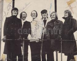 Az 1968-ban alapított The King's Singers brit a cappella együttes fotója a zenekar közel eredeti felállásában, az énekesek aláírásával. Készült 1975 körül. Vintázs, ezüst nagyítású felvétel, mérete: 202x254 mm. Az oxfordi King's College kórustagjainak baráti társasága az 1960-as évek közepén alapította kizárólag énekhangon alapuló zenei társulatát, melyet egykori iskolájuk iránt tisztelegve The King's Singersnek neveztek el. A kezdetektől hat énekesből álló énekkarban a mai napig változatlan a felállás, két kontratenor, egy tenor, két bariton és egy basszus szólam alkotja a zenekar egyedi, azonnal felismerhető, világszerte ünnepelt hangzását. Az alapítás óta eltelt több mint 50 évben természetesen cserélődtek a tagok, de a zenekar a mai napig a legünnepeltebb a cappella társulatnak számít. Aláírt fotónk 1975 körül készült, a zenekar közel eredeti felállásában: Simon Carrington (bariton, alapító tag), Alastair Hume (kontratenor, alapító tag), Brian Kay (basszus, alapító tag), Anthony Holt (bariton), Alastair Thompson (tenor, alapító tag), Nigel Perrin (kontratenor). Jó állapotú fénykép, egykori ragasztócsíkok nyomával, jobb oldalán enyhe gyűrődésnyommal.