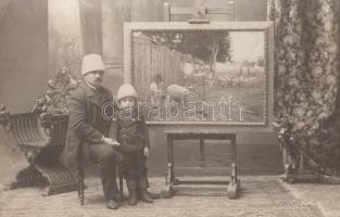 Oldal István festő és fotográfus nagybecskereki műtermében, kisfiával (1905.) Oldal István (1828-1916) nagybecskereki festő és fotográfus kucsmás, műtermében ülő, kettős portréja mellette álló (feltehetően saját) kisfiával, háttérben falusi zsánerfestményével, a lap hátoldalán ceruzával írt 1905-ös évszámmal. Vintázs, zselatinos ezüst nagyítás, mérete: 90x140 mm. A ,,nagybecskereki fényíró'', Oldal István 1829. augusztus 10-én született Szegeden, kőműves családban. 1853-ben kezdett fényképészettel foglalkozni, első műtermét is ekkor nyitotta meg a ,,Liptay kalaposmester temesvári utcai házának kapualjában''. Tanítómesterei valószínűleg vándorfényképészek voltak, akik a cirkuszokban, a majálisokon örökítették meg a polgárok, a szegényebb emberek arcképét. Az 1860-as évektől Oldal István már nem csak a műteremben készített felvételeket. Városokról és kultúrtörténeti eseményekről készített fotográfiáit, helytörténeti dokumentumait mindmáig féltve őrzik sok helyütt. Jó állapotú lap, hátoldalán megcímzéshez nyomtatott segédvonalakkal.