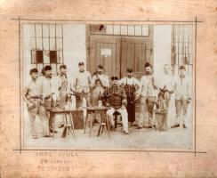 [Fotó] Szakaszvezető és kilenc kovácslegény beosztottja egy kecskeméti patkolóhelyiség bejárata előtt: Imre Gyula kecskeméti fotográfus felvétele. (1915 körül). Vintázs, zselatinos ezüst nagyítás, mérete, mérete: 110x150 mm egy 160x195 mm méretű kemény hátú fotókartonon. Első világháborús évekből származó kecskeméti felvételünk a karpaszományos szakaszvezetői egyenruhában ülő tiszt hátterében felsorakozott kilenc kovácslegényt ábrázolja, egy lovassági patkolóhelyiség bejárata előtt. Megkapó részlet, és komoly rangtiszteletről árulkodik a jelenség, miszerint az egység parancsnokának viaszkolt bajuszviselete valamennyi beosztottja arcán megjelenik, hol pörgébb, hol jelzésszerűbb változatban. A kilenc mesterlegényből hatan katonai zubbonyban sorakoztak fel, kilencük közül csupán egyetlen legény posztol feltűnően civil öltözetben, katonai csákó nélkül - a feltehetően inas pozíciójú civilt a kompozíció szélére száműzték marcona társai (vagy a hadi fotográfus) utasítására. A szakaszparancsnok közvetlen közelében, jobbra, kovácsmesteri kötényben a karpaszomány nélküli szakaszvezetői rangjelzést viselő műhelyvezető, a többi kovácslegény - a jelzés nélküli gallérhajtókából következően - közlegénynek minősül. Kecskeméti patkolóhelyiségünk földrajzi helyének azonosítása lehetetlen, a fénykép feltehetően a kecskeméti lovassági bázishoz tartozó kovácsműhely bejárata előtt készült. Fényképünk megkomponálója és készítője Imre Gyula fotográfus 1890-ben a kecskeméti iparos iskola fényképész tanonc hallgatója volt, és egészen az 1920-as évek derekáig a hirös városban működött. Kemény hátú fotókarton elhelyezett vintázs felvételünk körül kézi keretezés, alatta a fotográfus korabeli bélyegzése, a hátoldalon kézi feliratozás. A felvételen apró fakulások, a fotókartonon enyhe, halvány foltosság. Jó állapotú fotó és kartonlap.