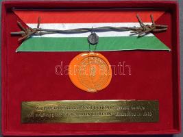 A Vasfüggöny egy darabja, plombálva, hitelesítő pecséttel, nemzeti színű szalag előtt, fémtáblán elhelyezett magyar nyelvű értelmező felirat kíséretében, díszdobozban. (1989). Történelmi emléknek számító tárgyi dokumentumunk az 1949-ben az aknazárral, szögesdróttal és őrtornyokkal kiépült Vasfüggöny egy darabja, mely osztrák oldalon 1955-ig, hazánkban egészen 1989-ig működött. A műszaki határzárat végül 1989 májusa és júliusa között távolította el a Magyar Néphadsereg. A történethez tartozó különös tény, hogy az osztrák és magyar külügyminiszter közös szimbolikus gesztusához, a vasfüggöny látványos átvágásához Sopron mellett újjáépítették az időközben már lebontott szögesdrót egy részét. A vasfüggöny zárótörténetének másik részét a maga tárgyiasságában hordozza tételünk: a szögesdrót keresett szuvenírré vált, melyet több különböző formában, díszes és kevésbé díszes kivitelben árusítottak. A nagyarányú hamisítások elkerülése érdekében a díszdobozos példányokhoz a Magyar Néphadsereg hitelesítő pecsétje és plombája, egyes esetekben sorszámozás is járt. (Példányunk sorszám nélküli.) A díszdoboz belsejében a 180 mm hosszúságú szögesdrót-darabka mellett gravírozott fémtábla értelmezi a műtárgyat. A díszdoboz fedelének belső oldalán az 1989. június 27-i vasfüggöny-átvágási ceremóniáról készített fényképfelvétel másolata, melyen Horn Gyula magyar és Alois Mock osztrák külügyminiszter közösen vágják át a határkerítést, magas rangú politikusok kíséretében. Tökéletes állapotú példány, vörös bársony bevonatú kartondobozban. Szép példány.