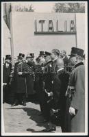 [Fénykép] Horthy Miklós kormányzó megtekinti a Budapesti Nemzetközi Vásárt, Chorin Ferenc titkos tanácsos társaságában. A Magyar Film Iroda felvétele. (1940). Vintázs, zselatinos ezüst nagyítás, mérete: 175x110, lapméret: 180x117 mm. A millenniumi kiállítás után időszerűvé vált, hogy Budapesten évente megrendezzék a hazai agrár- és ipari vállalkozásokat bemutató vásárt. A törekvések 1921-től értek be. A Horthy Miklós kormányzó jelenlétében megnyitott (ekkor még mezőgazdasági jellegű) vásár természetesen propagandacélokat is szolgált, a háborús veszteségből felálló ország gazdasági erejét volt hivatott hirdetni, ennek szellemében évente megrendezték a városligeti eseményt, egyre bővülő tematikával. Az 1930-as években már nemzetközi szinten jegyzett Budapesti Nemzetközi Vásár elnökévé Chorin Ferenc (1879-1964) titkos tanácsost választották, felvételünkön ő magyaráz a Horthy Miklós államfő vezette politikai testületnek. Felvételünk feltételezhetően 1940. április 26-án, pénteken, a vásár nyitónapján készült, Olaszország pavilonjánál. A kormányzó másik oldalán Bornemisza Géza volt iparügyi miniszter áll (1935-1939), a háttérben további politikusok, fejükön a legformálisabb viseletnek számító cilinderrel, a rendőrség díszszázadának tagja acél sisakban vigyázza a rendet. Fényképünk készülési idejét a jelenlevők listája, az olasz pavilon oldalán használt betűtípus és a kormányzó évről évre változó ruházata alapján azonosítottuk. Jó állapotú felvétel, hátoldalán jegyzetekkel.